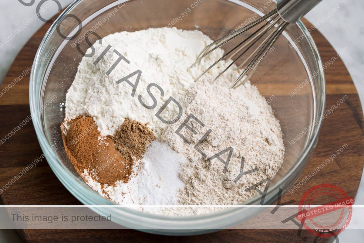 Mostrando cómo hacer muffins de campanilla. Batir la harina blanca, la harina de trigo, la canela, la nuez moscada, el bicarbonato de sodio y la sal en un bol de vidrio.